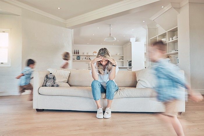 Frau verzweifelt auf Sofa zwei Kiner rennen um das Sofa herum.jpg